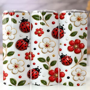 Puede incluir: Tres vasos blancos con un patrón repetido de mariquitas rojas, flores rojas y blancas, hojas verdes y detalles dorados. Los vasos tienen un acabado brillante y una tapa plateada.