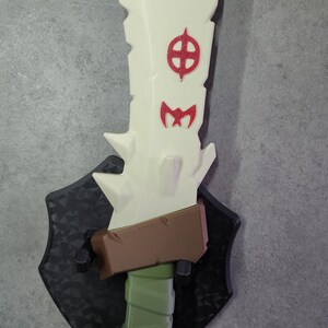 May include: A white jagged blade with red symbols mounted on a black shield-shaped wall plaque. The sword features a brown crossguard and a green wrapped handle. The blade has a rough, stone-like texture with sharp points along the side edges.