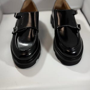 Könnte beinhalten: Ein Paar schwarze Lederschuhe mit glänzendem Finish. Jeder Schuh hat einen Riemen mit silberner Schnalle und eine dicke, strukturierte Sohle. Die Schuhe werden auf einer weißen Oberfläche präsentiert.