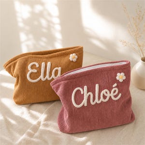 Pode incluir: Duas bolsas de cosméticos de veludo cotelê, uma amarelo mostarda e outra rosa empoeirado. Cada bolsa apresenta um nome bordado em branco, "Ella" e "Chloe", e um pequeno detalhe de margarida. As bolsas têm uma parte superior curva e um forro interno branco.
