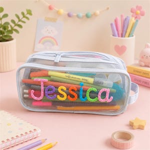 Op de afbeelding: Een lichtblauw etui van mesh met de naam Jessica in kleurrijke, gestructureerde, geborduurde letters op de voorkant. Het transparante etui is gevuld met diverse pennen en stiften en is voorzien van een handvat aan de zijkant.