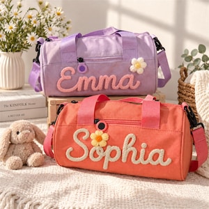 May include: Two cylindrical duffel bags, one lavender and one coral, each featuring a zippered front pocket, matching fabric handles, and a detachable shoulder strap. The lavender bag displays the name Emma, and the coral bag displays the name Sophia.