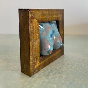 Op de afbeelding: Een klein, driedimensionaal keramisch beeldhouwwerk van een menselijke torso met een glanzend, gevlekt blauw en roodachtig roze glazuur. Het beeldhouwwerk is gemonteerd in een dikke, vierkante, donker gebeitste houten lijst.