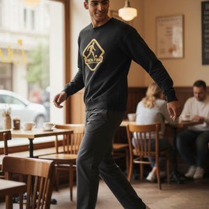 Puede incluir: Sudadera negra con un gráfico dorado de un jugador de fútbol y el texto "FINA FINA". La persona lleva pantalones grises oscuros y zapatillas blancas. La imagen está tomada en un café con mesas y sillas de madera.