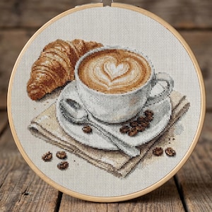 May include: Cross-stitch embroidery of a coffee cup with latte art in a heart shape, a croissant, coffee beans, and a spoon on a saucer. The embroidery is set in a wooden hoop, with a neutral-colored background.