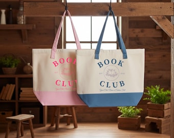 May include: Two canvas tote bags hanging from hooks. The left bag is cream with a pink base and pink text reading BOOK CLUB. The right bag is cream with a blue base and blue text reading BOOK CLUB. Both feature a central circular graphic design.