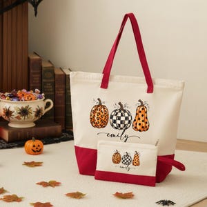 May include: A cream-colored canvas tote bag and matching zippered pouch with deep red trim. The front of both items features a graphic of three pumpkins with leopard print, checkered, and polka dot patterns, with the name emily written in script.