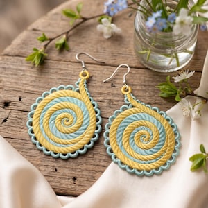 May include: A pair of dangle earrings featuring a circular embroidered design with a spiral pattern in alternating yellow and light blue thread. The edges are finished with a scalloped light blue border, and each earring hangs from a silver-toned hook.