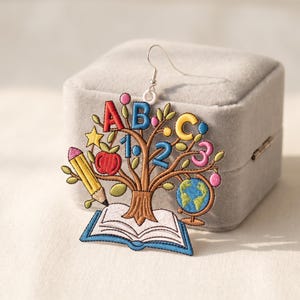 May include: A school-themed dangle earring featuring an embroidered tree design with a brown trunk rooted in an open book. The branches display colorful letters A, B, C, numbers 1, 2, 3, a yellow star, a red apple, a yellow pencil, and a globe.