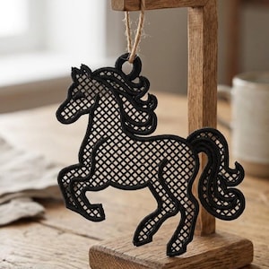 Puede incluir: Un adorno de caballo negro con un diseño calado detallado. El caballo está suspendido por una cuerda de hilo natural de un soporte de exhibición de madera. El adorno tiene una apariencia decorativa y elegante.