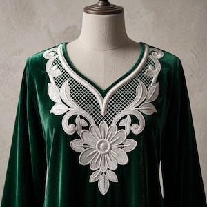 May include: A deep emerald green velvet garment featuring a V-neckline adorned with intricate white embroidery. The design includes a central floral motif surrounded by scrolling leaf patterns and a delicate grid-like lattice detail along the collar.