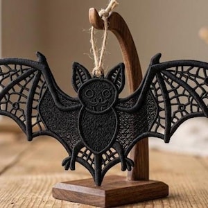 May include: A black embroidered bat ornament with outstretched wings, displayed on a wooden stand. The bat has a detailed design, with openwork wings and a smiling face. A natural twine loop is attached for hanging. The ornament is a Halloween or gothic-themed decoration.