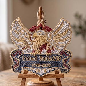 Pode incluir: Emblema patriótico bordado com uma águia dourada com as asas abertas. O design inclui uma faixa com "United States 250" e os anos "1776-2026". O emblema é exibido em um pequeno cavalete de madeira.
