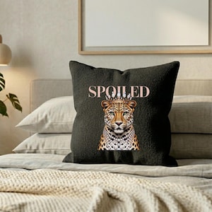 May include: A dark gray decorative throw pillow with the word "SPOILED" in pink lettering. The pillow features a regal leopard illustration wearing a crown and a pearl necklace.