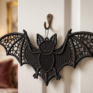 Op de afbeelding: Zwarte vleermuisvormige wanddecoratie met gedetailleerde vleugelpatronen, opgehangen aan een bronzen haak. De vleermuis heeft een getextureerde afwerking. Een decoratief item voor Halloween.