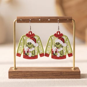 May include: A pair of dangling earrings shaped like miniature sweaters in lime green and red. The sweaters feature an embroidered green character wearing a red and white Santa hat, holding a white gift box with a bow, and hanging from silver hooks.