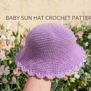 Op de afbeelding: Een lavendelkleurige gehaakte baby zonnehoed met een geschulpte rand. De tekst "BABY SUN HAT CROCHET PATTERN" staat boven de hoed. De hoed wordt getoond tegen een achtergrond van bloemen.