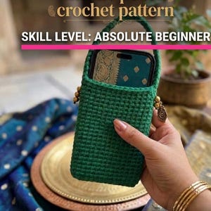 Puede incluir: Una funda de ganchillo verde para móvil con asa, que sostiene un teléfono. El texto de la imagen dice "mobile pouch crochet pattern" y "SKILL LEVEL: ABSOLUTE BEGINNER". La funda está decorada con cuentas doradas y un dije de pluma. El texto inferior dice "A ONE-DAY CROCHET PROJECT FOR TOTAL BEGINNERS."