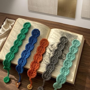 May include: Five crochet bookmarks in various colours lie on an open book. The bookmarks are green, blue, orange, grey, and teal. Each has a button or wooden charm. The book is open to a page with printed text and illustrations.