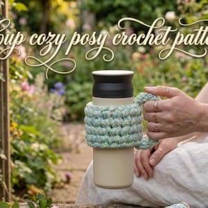 Op de afbeelding: Een beige reismok met een zwart deksel en een lichtblauwe en groene gehaakte hoes. De hoes heeft een handvat en de tekst "Cup cozy posy crochet pattern" in een decoratief lettertype. De achtergrond is een tuin.