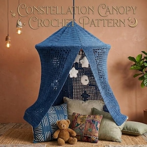 May include: A blue crochet canopy with the text "Constellation Canopy Crochet Pattern" hangs above pillows and a teddy bear. The canopy is suspended from the ceiling and has a star and moon design. The pillows are various colors and patterns.