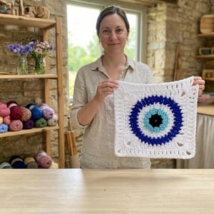 May include: A white crochet square featuring a blue and black eye design. The square is held up in front of a person. The background includes shelves with yarn and wooden items, and a window.