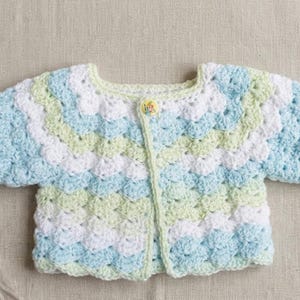 May include: A handmade, crocheted baby cardigan in a shell stitch pattern. The sweater features a color scheme of light blue, white, and pale green. It has a single button closure and is designed for a baby or toddler.