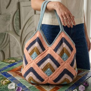 Può includere: Una borsa tote all'uncinetto color pesca, blu e blu navy con un motivo geometrico. La borsa ha un'apertura a forma di V e manici azzurri. La borsa è appoggiata su un tavolo colorato.