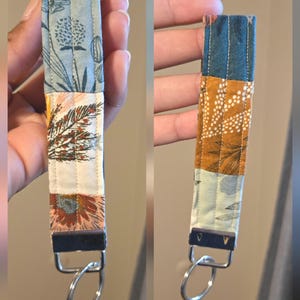 May include: A fabric wristlet keychain featuring a patchwork design with floral and botanical patterns in shades of blue, orange, white, and teal. The strap is secured with a dark metal clamp and includes a circular silver-toned metal key ring.