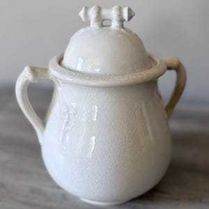 May include: A white ceramic mug with a black handle and the text "But First, Coffee" in a black, handwritten-style font. The mug is on a white surface.