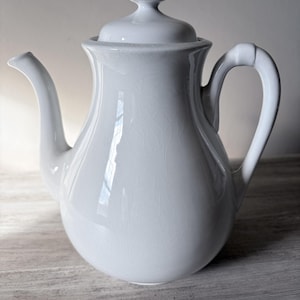May include: A white ceramic teapot with a lid and a curved handle. The teapot has a bulbous body and a spout. The lid is topped with a decorative finial. The teapot is sitting on a wooden surface.