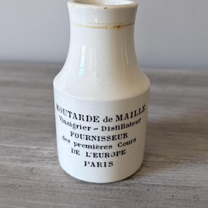 May include: A white ceramic bottle with a slightly flared shape. The bottle has black text that reads "Moutarde de Maille, Vinaigrier - Distillateur, Fournisseur des premières Cours DE L'EUROPE PARIS". The top of the bottle has a gold band.