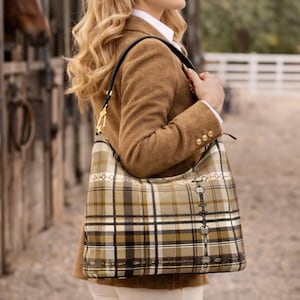 May include: A brown plaid shoulder bag with a black strap and gold hardware. The bag features a pattern of tan, black, and white squares. The bag is carried by a person wearing a brown blazer and white trousers. The background includes a horse stable.