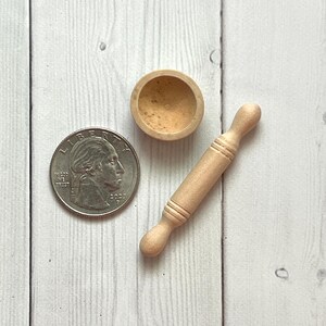 May include: A miniature wooden rolling pin, a small bowl, and a U.S. quarter coin are arranged on a light-coloured wooden surface. The rolling pin is approximately 7.6 cm long. The bowl is round and contains a light-brown substance.
