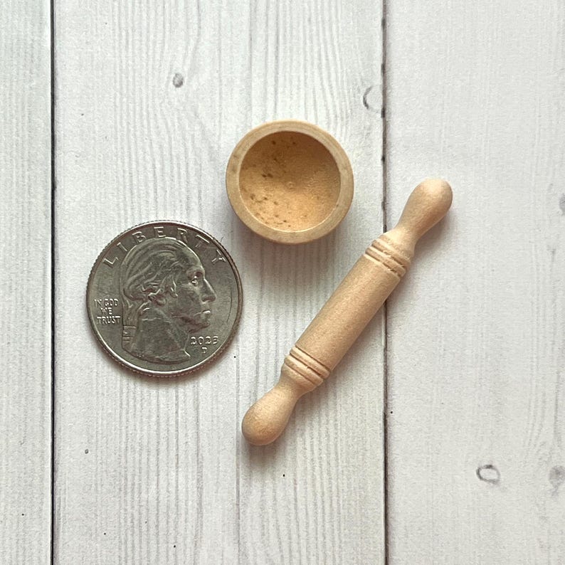 May include: A miniature wooden rolling pin, a small bowl, and a U.S. quarter coin are arranged on a light-coloured wooden surface. The rolling pin is approximately 7.6 cm long. The bowl is round and contains a light-brown substance.