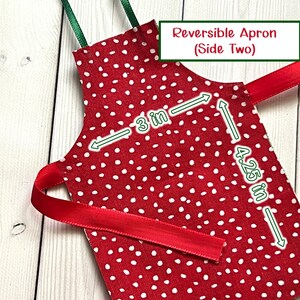 May include: A red and white polka dot reversible apron with green and red ribbon ties. The apron measures 7.6 cm by 10.8 cm. The text "Reversible Apron (Side Two)" is in a green and white box.