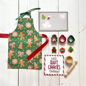 May include: A festive green apron featuring gingerbread men and candy canes, alongside cookie cutters, a rolling pin, a small wooden bowl, and a sign saying "Let's BAKE COOKIES today!" in red and white.