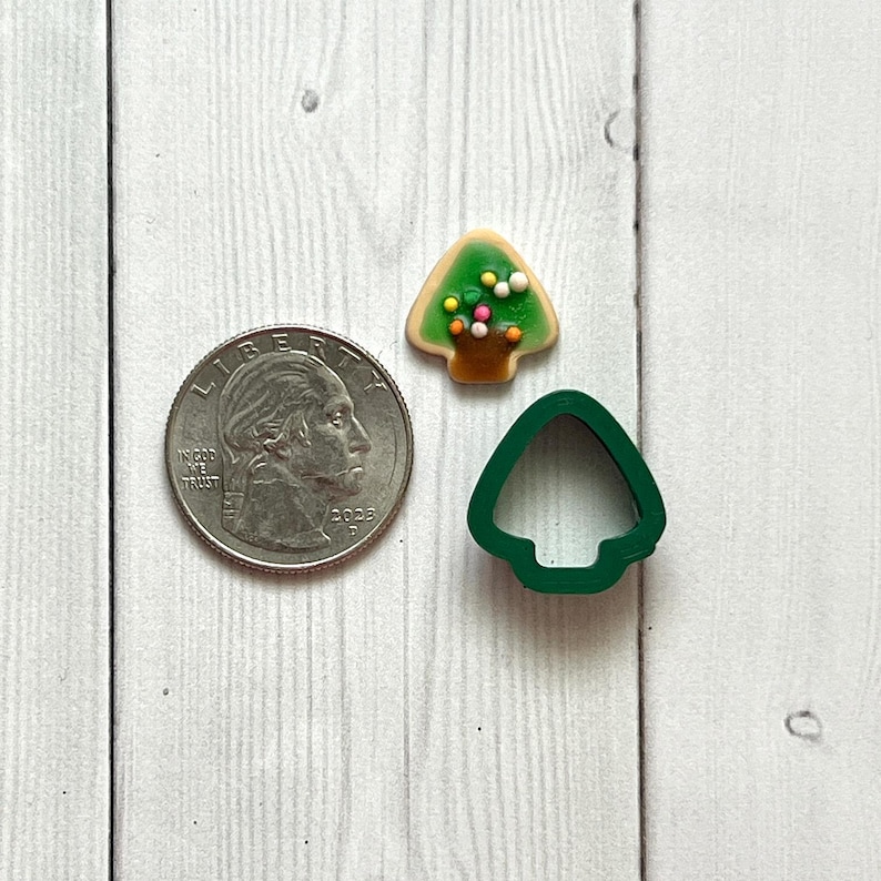 May include: A US quarter next to a decorated Christmas tree biscuit and a green tree-shaped biscuit cutter. The biscuit has green icing with colourful sprinkles and a brown trunk. The cutter is made of green plastic.