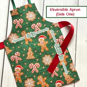 May include: Reversible apron with a festive Christmas pattern. The green fabric features gingerbread men, candy canes, and snowflakes. The apron is 7.6 cm wide and 10.8 cm long. The text on the apron says "Reversible Apron (Side One)".