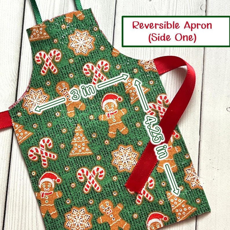 May include: Reversible apron with a festive Christmas pattern. The green fabric features gingerbread men, candy canes, and snowflakes. The apron is 7.6 cm wide and 10.8 cm long. The text on the apron says "Reversible Apron (Side One)".