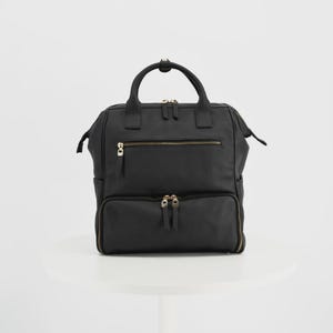 Könnte beinhalten: Ein schwarzer Rucksack mit einem Tragegriff, zwei Schultergurten und einem Hauptfach mit Reißverschluss. Die Vorderseite verfügt über eine horizontale Reißverschlusstasche und eine größere untere Reißverschlusstasche mit goldfarbenen Beschlägen.