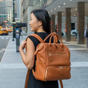 Può includere: Uno zaino in pelle marrone con cerniere e manici dorati. La borsa ha più scomparti e viene indossata da una persona su un marciapiede cittadino. Lo zaino ha un design strutturato e un aspetto classico.