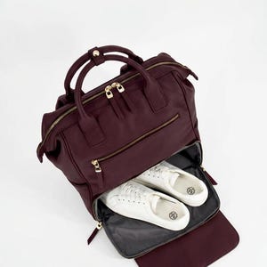 Könnte beinhalten: Ein tiefburgunderfarbener Lederrucksack mit goldfarbenen Reißverschlüssen und Tragegriffen. Das vordere Reißverschlussfach enthält ein Paar weiße Low-Top-Sneaker mit Schnürsenkeln und zeigt ein funktionales Design zur Aufbewahrung.
