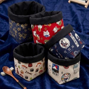 May include: Five small fabric storage baskets with black rims. The baskets feature various Japanese-themed patterns, including floral, cat, sushi, and landscape designs. A wooden spoon rests on a small white holder, adding a decorative touch to the scene.