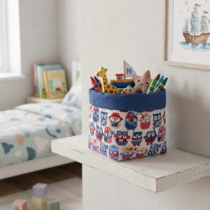 May include: A fabric storage bin with a blue rim and owl pattern, filled with toys and crayons. The bin sits on a white shelf in a child's room. Toys include a giraffe, boat, and star.