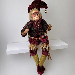 May include: A decorative monkey doll wearing a deep burgundy velvet jester hat with gold star accents and a matching bell. The doll is dressed in a dark patterned jacket, burgundy velvet pants with gold fringe trim, and gold glittery leggings.