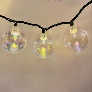 May include: String of clear, spherical fairy lights with a shimmering, iridescent effect. Each bulb is lit, emitting a warm glow. The lights are strung on a black wire, suitable for indoor or outdoor decor.