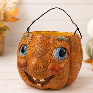 May include: A whimsical orange pumpkin-shaped container with a smiling face, blue eyes, and a black handle. The container has a textured surface and is likely used for Halloween decorations or as a candy holder.