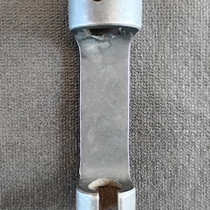 May include: A silver-toned metal mechanical component featuring a cylindrical top section with a circular hole, a flat rectangular central body, and a bottom section with a U-shaped cutout. The surface shows a matte finish with visible wear.