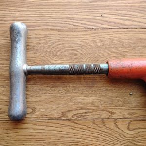 Pode incluir: Uma ferramenta de metal antiga com uma al&ccedil;a em forma de T e uma pega laranja. A al&ccedil;a &eacute; prateada e mostra sinais de desgaste. A pega laranja tem um design curvo. A ferramenta est&aacute; sobre um fundo de madeira.